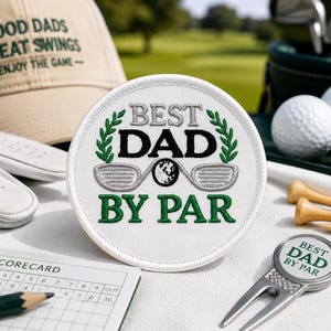 Puede incluir: Un parche bordado blanco de forma circular con el texto BEST DAD BY PAR en hilo negro y verde, con una pelota de golf y dos palos de golf bordados en el centro, rodeados de hojas de laurel verdes y un borde blanco cosido.