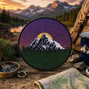May include: A circular embroidered patch featuring a mountain range with snow-capped peaks, a yellow sun, and a dark green forest foreground. The sky is depicted with horizontal lines in shades of purple and orange, enclosed by a black stitched border.