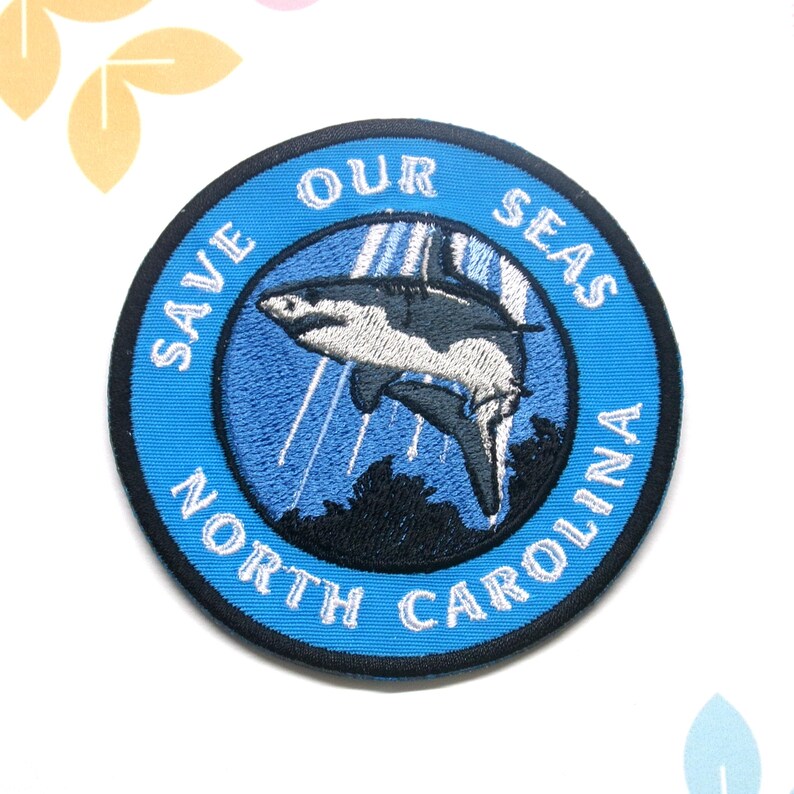 May include: Embroidered patch with a blue border and the text "SAVE OUR SEAS NORTH CAROLINA." The patch features a detailed illustration of a shark in shades of gray and white, set against a blue background.