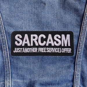 May include: A black rectangular patch with the word "SARCASTM" embroidered in white, above the text "JUST ANOTHER FREE SERVICE I OFFER". The patch is sewn onto a blue denim fabric.