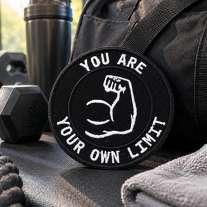Puede incluir: Un parche bordado negro de forma circular que presenta el contorno blanco de un brazo con el bíceps flexionado en el centro. El texto YOU ARE YOUR OWN LIMIT está bordado en letras mayúsculas blancas alrededor del borde del diseño circular.