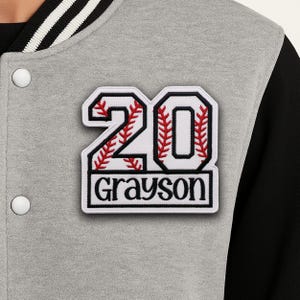 May include: A grey letterman jacket with a black sleeve and white and black striped trim. The jacket features a white patch with the number "20" in a baseball stitch design and the name "Grayson" below.