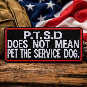 Puede incluir: Un parche rectangular negro con texto blanco que dice "P.T.S.D DOES NOT MEAN PET THE SERVICE DOG." El parche tiene un borde rojo. El fondo incluye una bandera estadounidense y una bolsa de estilo militar.