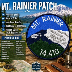 May include: A circular embroidered patch featuring a blue sky, a dark blue mountain silhouette, a white snow-capped peak, and green forest base. The text reads Mt. Rainier and 14,410 in white thread. The patch has a dark blue stitched border.