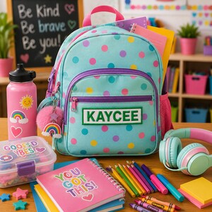 K&ouml;nnte beinhalten: Ein hellblauer Rucksack mit bunten Punkten und einem rechteckigen Aufn&auml;her mit der Aufschrift KAYCEE. Der Rucksack hat einen rosa Tragegriff, violette Rei&szlig;verschl&uuml;sse und einen rosa Bommel-Schl&uuml;sselanh&auml;nger, umgeben von Trinkflasche und Heften.