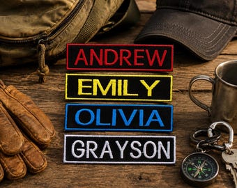 Könnte beinhalten: Vier rechteckige gestickte Aufnäher mit schwarzem Hintergrund und Rand. Der oberste Aufnäher zeigt den Namen ANDREW in Rot, der zweite EMILY in Gelb, der dritte OLIVIA in Blau und der unterste Aufnäher zeigt den Namen GRAYSON in Weiß.