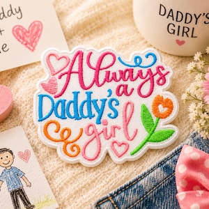 Puede incluir: Un parche bordado blanco con el texto Always a Daddy's girl en letras cursivas y de molde rosas y azules. El diseño incluye un pequeño tulipán naranja con un centro en forma de corazón, hojas verdes y varios contornos de corazones rosas.