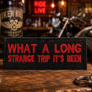 May include: A rectangular black embroidered patch featuring the red stitched text "WHAT A LONG STRANGE TRIP IT'S BEEN" in a bold, capitalized font. The patch has a clean, finished border and is designed for attachment to clothing or gear.
