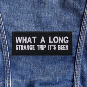 Peut inclure: Un écusson rectangulaire noir avec le texte brodé en blanc "WHAT A LONG STRANGE TRIP IT'S BEEN." L'écusson est cousu sur un tissu en denim bleu, probablement une veste ou un gilet.