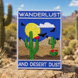 May include: A rectangular embroidered patch featuring a blue background with the text WANDERLUST at the top and AND DESERT DUST at the bottom. The design shows a yellow sun, a white cloud, black birds, and green cacti with red flowers.