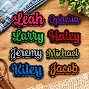 Op de afbeelding: Een verzameling geborduurde naamlabels in diverse kleuren, waaronder rood, groen, paars en oranje. Elk label toont een andere naam in een gestileerd lettertype met een zwarte rand. De namen zijn Leah, Ophelia, Larry, Haley, Jeremy, Michael, Kiley en Jacob.