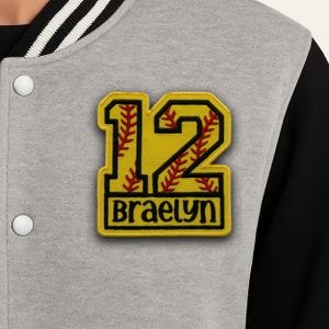 Può includere: Giacca college grigia e nera con una toppa gialla con il numero 12 e il nome "Braelyn" in lettere nere. Il numero 12 ha cuciture rosse e la giacca ha dettagli a righe bianche e nere.