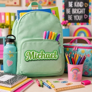 K&ouml;nnte beinhalten: Ein hellgr&uuml;ner Rucksack mit dem Namen Michael in wei&szlig;er Schreibschrift auf der Vordertasche. Der Rucksack hat eine seitliche Netztasche mit einer hellgr&uuml;nen Trinkflasche mit Herz- und Sternaufklebern sowie sechs bunte Filzstifte vorne.