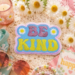 May include: A fabric patch with a light blue background and a scalloped lavender border. The word BE is embroidered in purple, and the word KIND is embroidered in lime green. Two small white daisies with yellow centers flank the word BE.