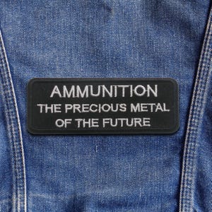 Könnte beinhalten: Ein schwarzer rechteckiger Aufnäher mit weiß gesticktem Text: „AMMUNITION THE PRECIOUS METAL OF THE FUTURE“. Der Aufnäher ist auf einem blauen Jeanskleidungsstück befestigt. Der Aufnäher hat einen schwarzen Rand.