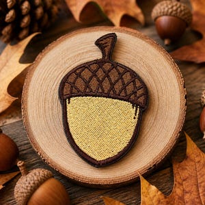 May include: An acorn-shaped embroidered patch in brown and yellow, displayed on a wooden slice. The patch has a detailed, textured top and a smooth, yellow base. Acorns and autumn leaves are scattered around the patch.