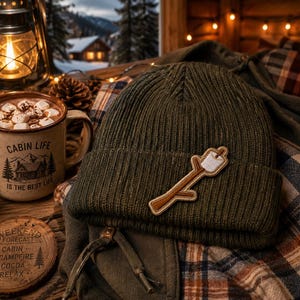 May include: An olive green knit beanie with a marshmallow on a stick patch rests on a plaid shirt. A mug of hot chocolate with marshmallows sits next to a wooden coaster that reads "Weekend Forecast: Cabin, Campfire, Cocoa, Relax."