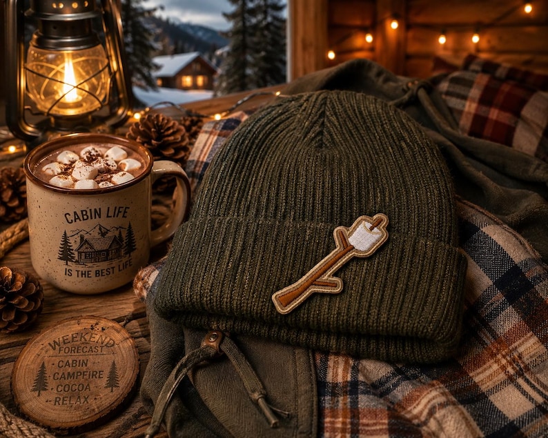 May include: An olive green knit beanie with a marshmallow on a stick patch rests on a plaid shirt. A mug of hot chocolate with marshmallows sits next to a wooden coaster that reads "Weekend Forecast: Cabin, Campfire, Cocoa, Relax."