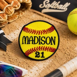 May include: A yellow softball patch with the name "MADISON" and the number "21" in black lettering, surrounded by red stitching. The patch is on a sandy surface, with a softball, glove, sunglasses, and a bow.