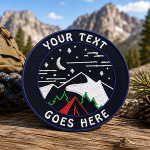 May include: A circular embroidered patch with a dark blue background featuring a white crescent moon, stars, and mountain peaks above green pine trees and a red tent. The text "YOUR TEXT GOES HERE" is stitched in white along the top and bottom edges.