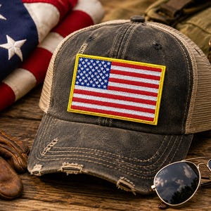 May include: A distressed charcoal grey baseball cap featuring a rectangular embroidered patch of the United States flag with a yellow border. The cap has a mesh back panel and a curved brim with frayed edges, showcasing a worn-in aesthetic.