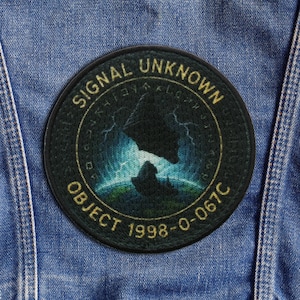 May include: A circular, dark green patch with gold text reading "SIGNAL UNKNOWN" and "OBJECT 1998-O-067C". The patch depicts a mountain silhouette against a blue and green backdrop with lightning.