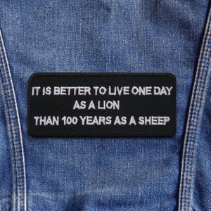Op de afbeelding: Een zwart rechthoekig embleem met wit geborduurde tekst. De tekst luidt: "HET IS BETER OM EEN DAG ALS LEEUW TE LEVEN DAN 100 JAAR ALS SCHAAP." De patch is op een blauwe denim stof genaaid.