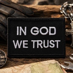 May include: A rectangular black patch featuring the white embroidered text IN GOD WE TRUST in a bold, sans-serif font. The patch has a stitched border and a flat, fabric-like texture, presenting a clear contrast between the dark background and white lettering.