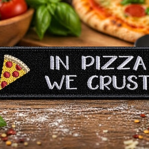 Puede incluir: Parche negro con texto bordado en blanco "IN PIZZA WE CRUST" y un gráfico de una porción de pizza. La porción de pizza tiene una corteza amarilla, pepperoni rojo y un borde blanco. El parche está sobre una superficie de madera con pizza e ingredientes.
