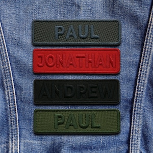 May include: Four rectangular embroidered name patches: "PAUL", "JONATHAN", "ANDREW", and "PAUL". The patches are in green, red, and black, with raised, textured lettering. The patches are set against a blue denim background.