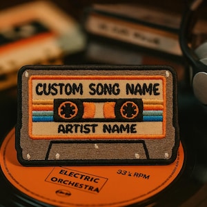 Op de afbeelding: Geborduurde patch in de vorm van een cassettebandje met de tekst "CUSTOM SONG NAME" en "ARTIST NAME". De patch heeft oranje, blauwe en bruine strepen. Het ligt op een oranje plaatlabel met de tekst "ELECTRIC ORCHESTRA".