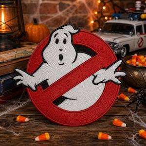 May include: A circular embroidered patch featuring a white ghost character with black outlines and facial features, centered within a red circle with a diagonal red slash across it, mimicking the iconic Ghostbusters logo design.