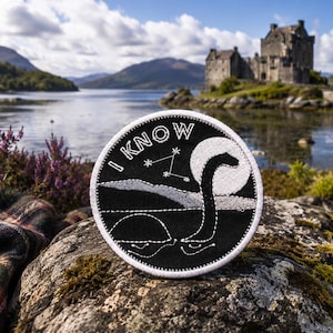 May include: A round, black and white embroidered patch featuring the Loch Ness Monster with the text "I KNOW". The design includes a full moon, stars, and a stylized depiction of the famous cryptid. The patch is set against a scenic backdrop of a lake and castle.