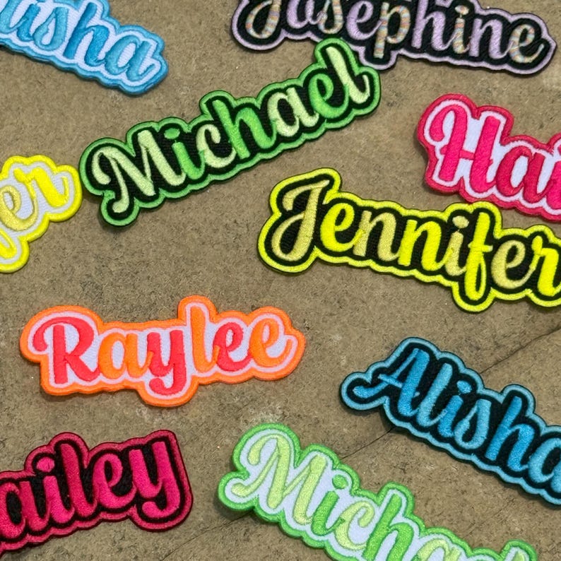 K&ouml;nnte beinhalten: Eine Sammlung von bestickten Namensaufn&auml;hern in verschiedenen Farben, darunter Neongr&uuml;n, leuchtendes Pink, Gelb, Orange und Hellblau. Jeder Aufn&auml;her zeigt eine Schreibschrift mit einem dicken Rand und Namen wie Michael, Jennifer, Raylee und Alisha.