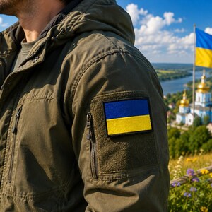 May include: An olive green jacket with a Ukrainian flag patch on the sleeve. The jacket has a hood and multiple pockets. In the background, a Ukrainian flag waves in the wind, with a church and landscape visible.
