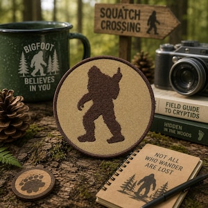 May include: A circular embroidered patch featuring a dark brown silhouette of Bigfoot on a tan background with a dark brown border. The patch is displayed alongside a green mug, a notebook, a camera, and a wooden sign with Bigfoot imagery.