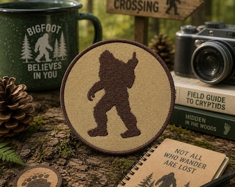 May include: A circular embroidered patch featuring a dark brown silhouette of Bigfoot on a tan background with a dark brown border. The patch is displayed alongside a green mug, a notebook, a camera, and a wooden sign with Bigfoot imagery.