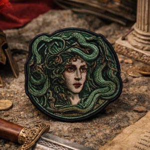 May include: A circular embroidered patch featuring a detailed portrait of Medusa with green snakes intertwined in her hair. The design uses shades of green, brown, and cream thread against a dark background, enclosed by a black stitched border.