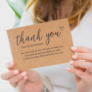 May include: A brown card with the words "thank you" in elegant script, along with the message "FOR YOUR ORDER." A small heart is above the word "you." The card is held by a person with light pink nail polish.