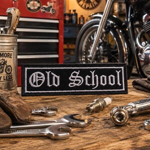 May include: A black rectangular patch with the words "Old School" embroidered in white script. The patch is on a wooden surface with tools, gloves, and a motorcycle in the background. The scene evokes a vintage garage or workshop aesthetic.
