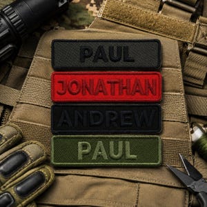 Puede incluir: Cuatro parches rectangulares bordados con cierre de velcro. El parche superior es negro con el nombre PAUL en hilo negro. El segundo es rojo con JONATHAN en rojo. El tercero es negro con ANDREW en negro. El inferior es verde oliva con PAUL en verde oliva.