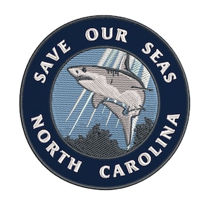 May include: Embroidered patch with a navy blue border and the text "SAVE OUR SEAS NORTH CAROLINA" in white. The patch features a shark leaping out of the water, with light blue and gray accents. The design is circular.