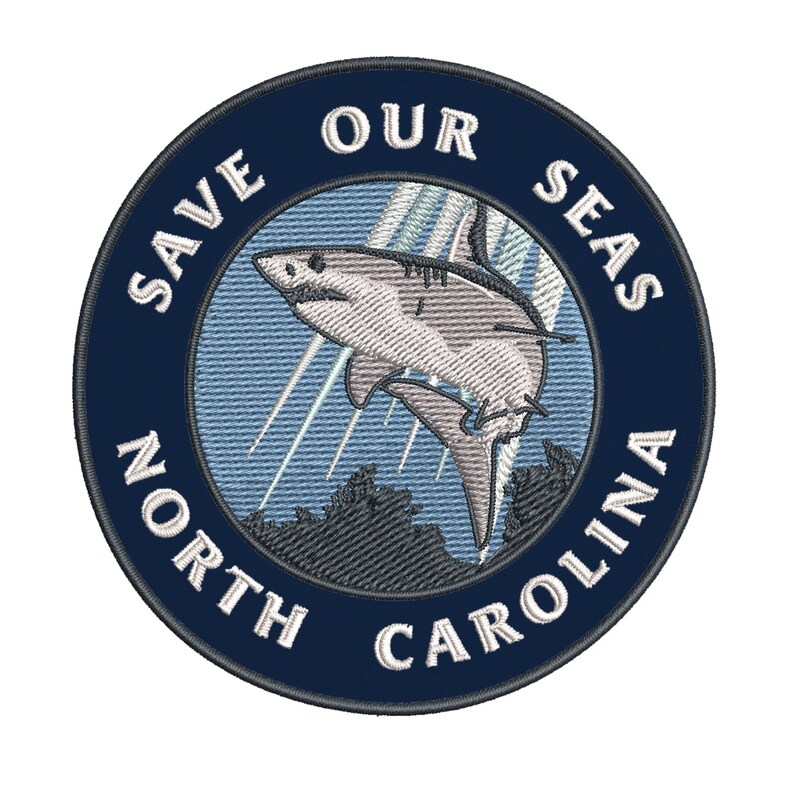 May include: Embroidered patch with a navy blue border and the text "SAVE OUR SEAS NORTH CAROLINA" in white. The patch features a shark leaping out of the water, with light blue and gray accents. The design is circular.