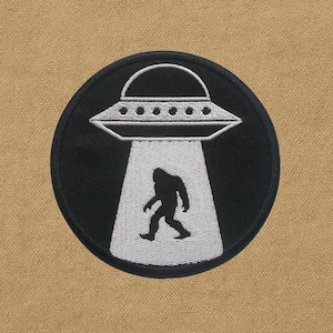 Peut inclure: Un écusson rond noir avec un dessin d'OVNI blanc, projetant un faisceau lumineux sur une silhouette de Bigfoot. L'OVNI a de petites lumières espacées. Le Bigfoot marche vers l'avant. L'écusson est sur un fond beige.