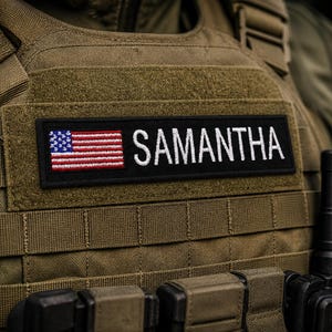 May include: A close-up shot of a tactical vest with a name patch. The patch displays the name "SAMANTHA" in white capital letters, alongside a small American flag. The vest is olive green and has multiple straps and pockets.