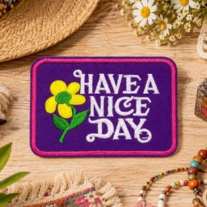 May include: A rectangular embroidered patch with a purple background and a pink border. The patch features a yellow flower with a green stem and the white text "HAVE A NICE DAY" with a small smiley face integrated into the letter Y.