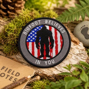 Pode incluir: Remendo bordado circular com uma silhueta de Bigfoot contra um desenho de bandeira americana vermelha, branca e azul. O remendo diz "BIGFOOT BELIEVES IN YOU". O remendo está em uma superfície de madeira, cercado por musgo e folhagem.