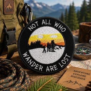 May include: A circular embroidered patch with a black border featuring the white text NOT ALL WHO WANDER ARE LOST. The center depicts a landscape with a white snowy foreground, black silhouettes of trees and people, and a yellow and orange sky.