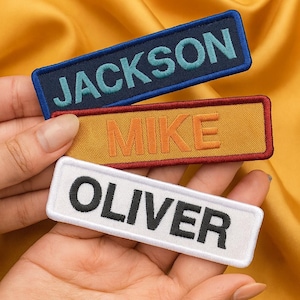 May include: Three embroidered name patches with the names JACKSON, MIKE, and OLIVER. The JACKSON patch is navy blue with teal lettering, the MIKE patch is gold with orange lettering, and the OLIVER patch is white with black lettering. The patches are rectangular.