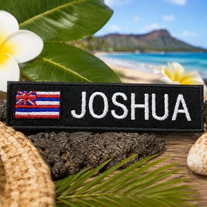 May include: A rectangular black embroidered patch featuring the name JOSHUA in white capital letters next to a small, detailed representation of the Hawaii state flag with its red, white, and blue horizontal stripes and canton design.